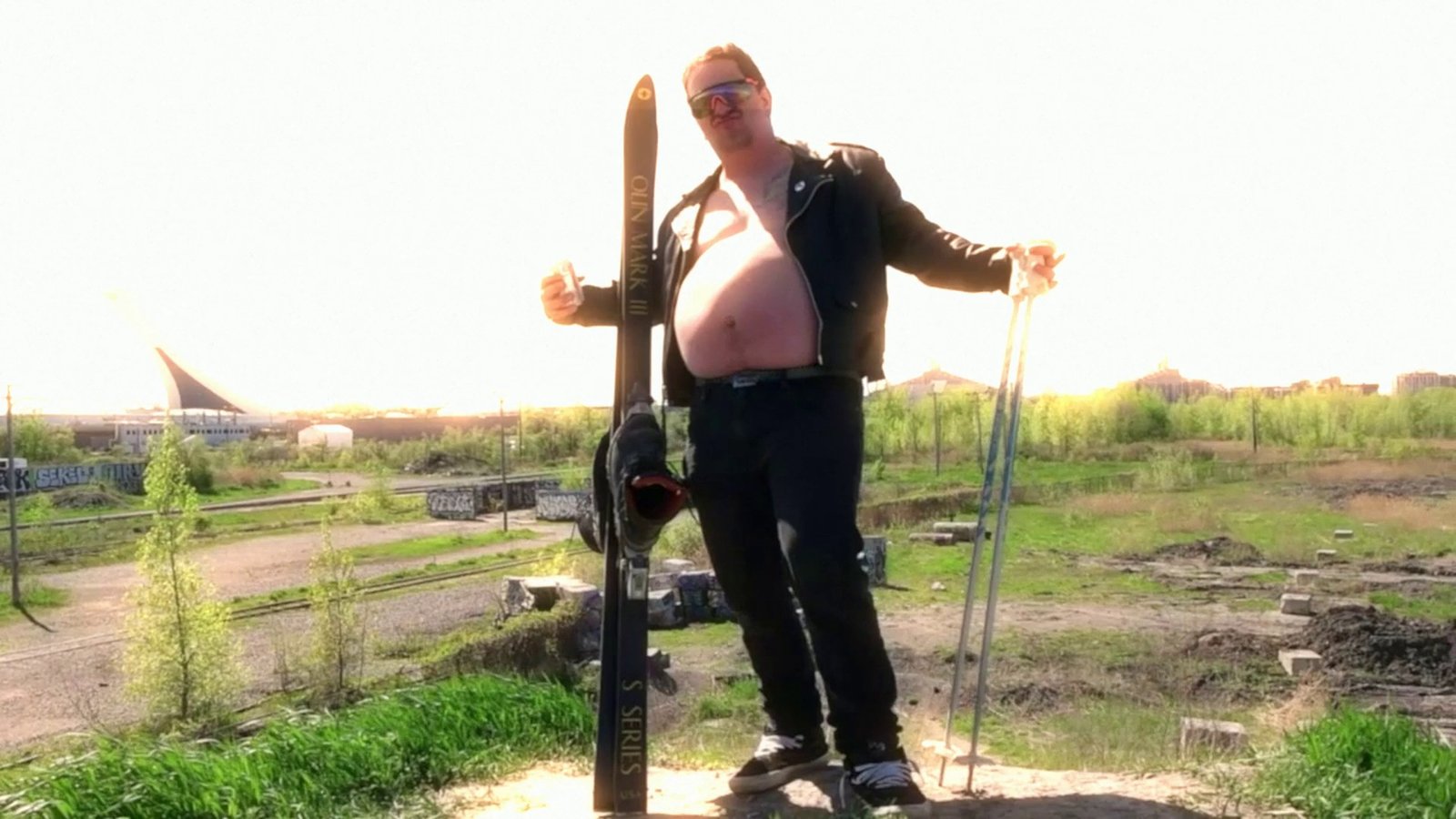 Daylight footage: a skier posing on a dirt lot with a stadium behind. After Super8 Film Lab, halation look: halation at 200 percent, bloom low.