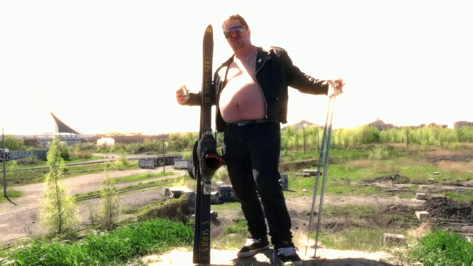 Daylight footage: a skier posing on a dirt lot with a stadium behind. After Super8 Film Lab, glow look: bloom at 180 percent with strong halation.