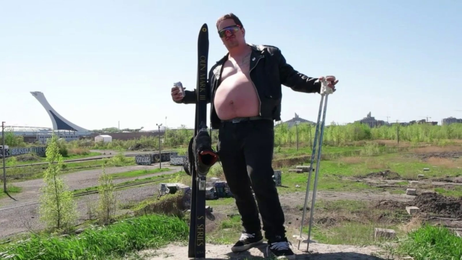 Source frame, untouched: Daylight footage: a skier posing on a dirt lot with a stadium behind.
